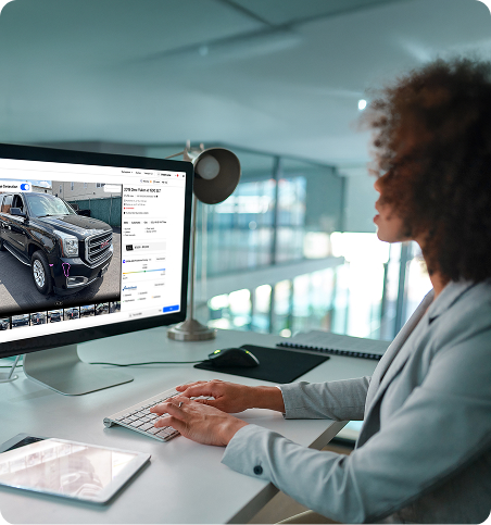 A person with curly hair sits at a desk using a computer to browse an online car marketplace, viewing a listing for a black pickup truck. A tablet and desk lamp are also on the desk.