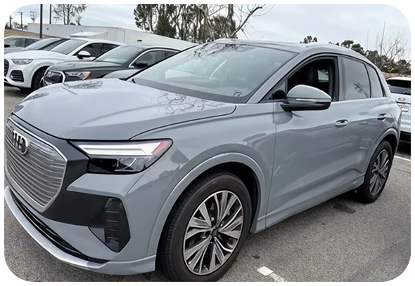 A silver Audi electric SUV is parked in a lot with other cars. The vehicle features modern styling, sharp headlights, and a prominent front grille. Trees and a cloudy sky are visible in the background.