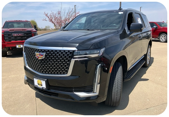 A black Cadillac Escalade SUV is parked outdoors on a sunny day, with a red GMC vehicle visible in the background. The Escalade is facing slightly left, showcasing its front grille and sleek design.
