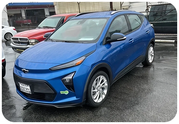 A blue Chevrolet Bolt EUV electric car is parked on a wet pavement near other vehicles on a rainy day, with a license plate visible on the front.