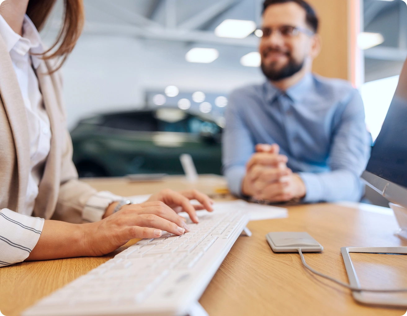 A person types on a white keyboard at a desk while another person sits across, smiling, in an office setting with a blurred car visible in the background.