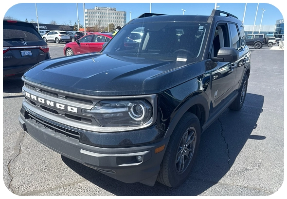 A black Ford Bronco SUV is parked in a lot on a sunny day, surrounded by other vehicles with buildings visible in the background.