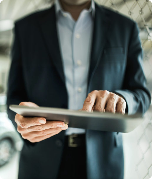 A person wearing a dark suit and white shirt uses a tablet, standing indoors in front of a chain-link fence, with the focus on their hands and the device.