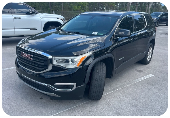 A black GMC SUV is parked in a parking lot with its front wheels turned outward. Other vehicles and trees are visible in the background.