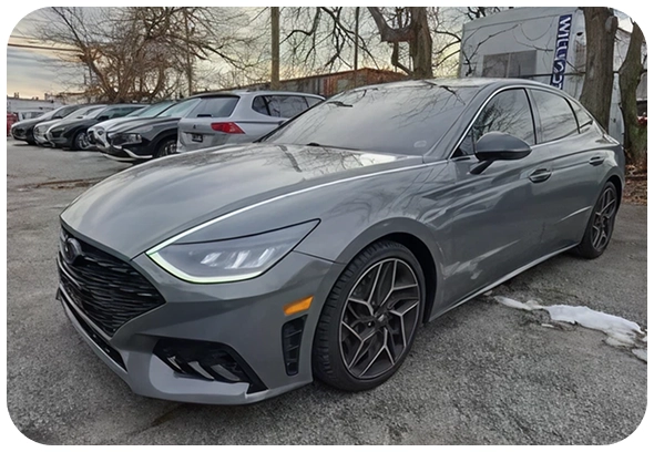 A sleek, gray sedan with modern styling is parked in a lot next to several other vehicles on a paved surface, with leafless trees and a building visible in the background.
