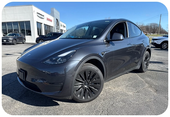 A dark gray Tesla Model Y parked outside a dealership on a sunny day, with other vehicles and a modern building visible in the background.