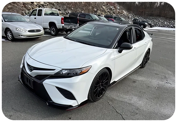 A white Toyota Camry sedan with black wheels and accents is parked in a lot with other cars and rocky terrain in the background.