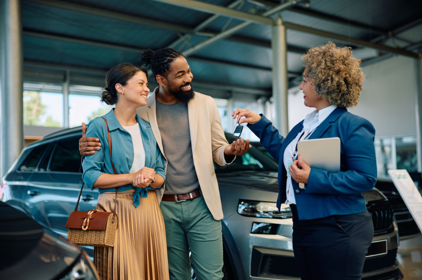 A smiling couple stands together as a car salesperson hands them a set of keys at a dealership. The couple looks happy, and new cars are visible in the background.