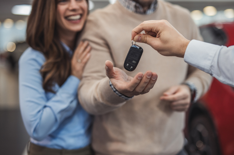 A person hands car keys to a smiling couple in a dealership. The man reaches out to accept the keys while the woman stands beside him, looking happy and excited—enjoying buyer protection for added peace of mind.