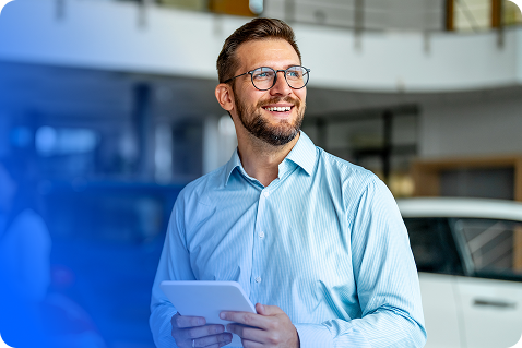 A smiling man with glasses and a beard stands indoors, holding a tablet and wearing a light blue shirt. There are blurred cars and bright indoor lighting in the background.