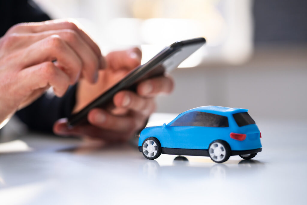 A person uses a smartphone near a small blue toy car on a white table, suggesting automotive apps.