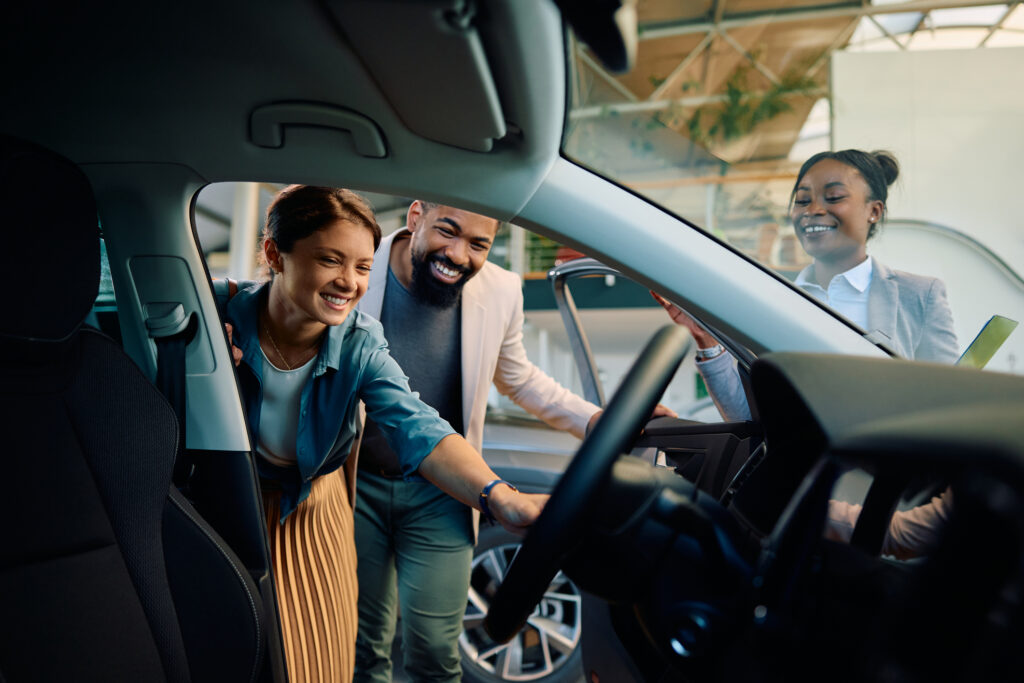 Three people smile and look inside a car, with one woman sitting in the driver’s seat and two others standing outside, suggesting they are at a car dealership.