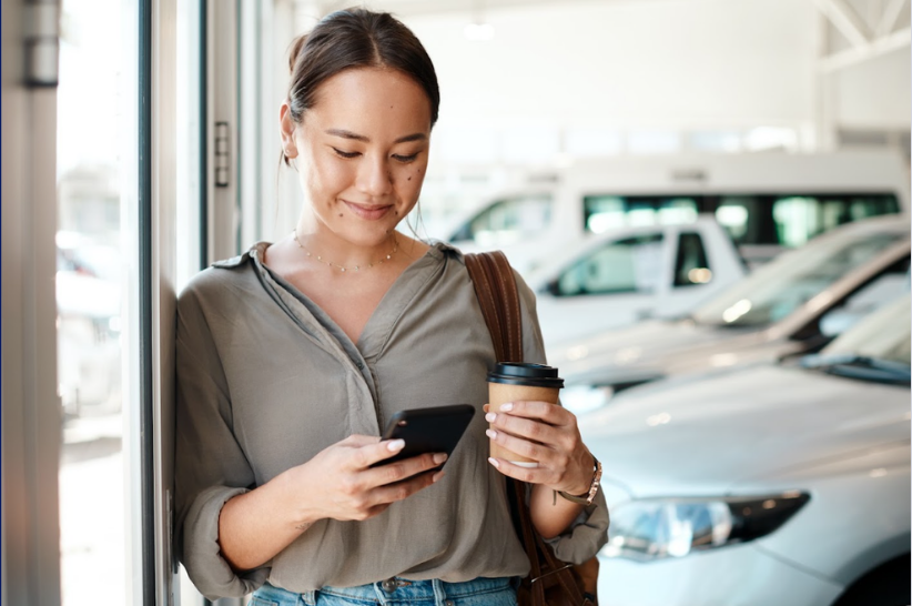 A woman stands indoors by cars, holding a coffee cup and looking at her smartphone, smiling. She wears a gray blouse and a brown bag over her shoulder, perhaps checking updates about vehicle arbitration as she enjoys her day.