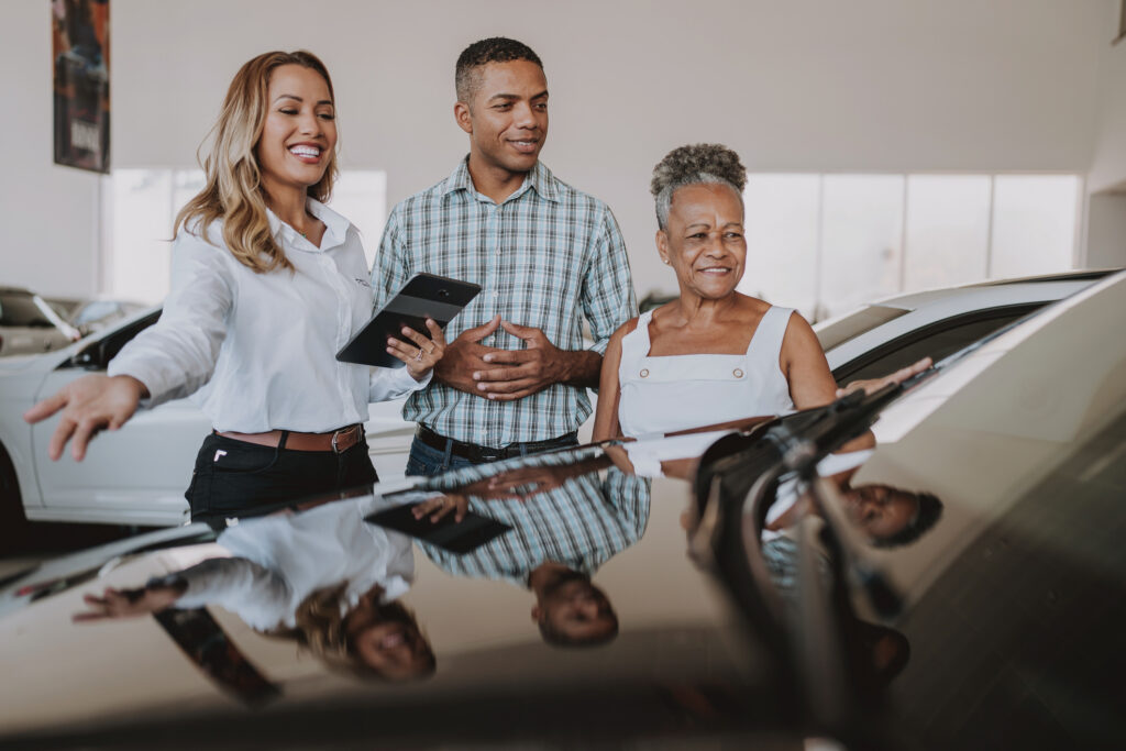 A smiling car salesperson shows a vehicle to a man and an older woman in a bright showroom. All three appear engaged and happy while discussing the car.