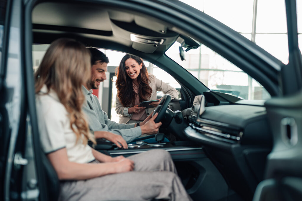 A man and woman sit in a car at a dealership, with a smiling saleswoman standing outside the drivers door, holding a clipboard and talking to them.