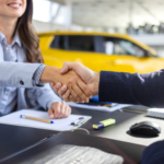 Two people in business attire shake hands across a desk with office supplies. A yellow car is visible in the background, suggesting a car dealership setting.