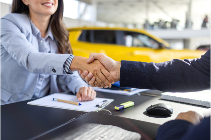 Two people in business attire shake hands across a desk with office supplies. A yellow car is visible in the background, suggesting a car dealership setting.