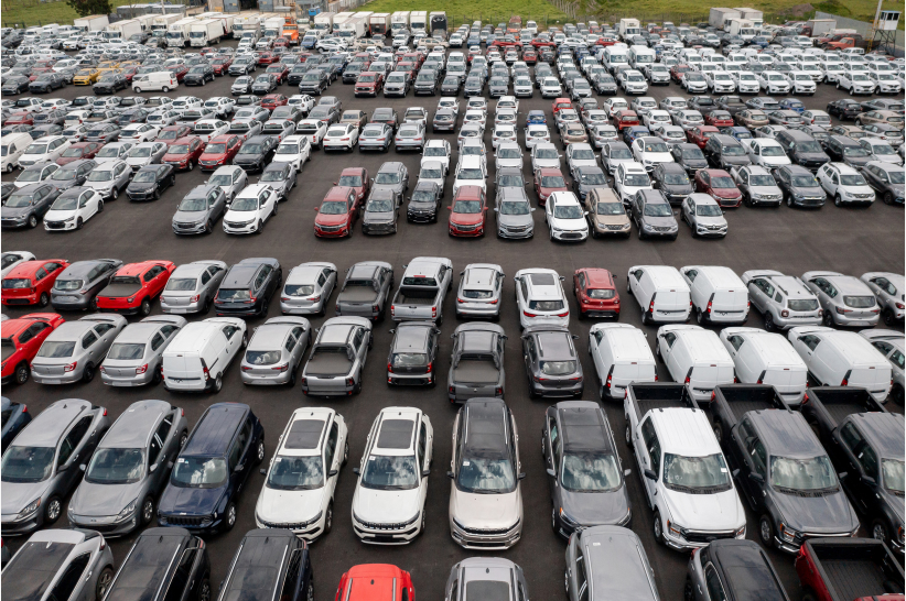 Hundreds of parked cars, mostly white and gray, fill a large outdoor lot. Vehicles are arranged in organized rows, viewed from above.