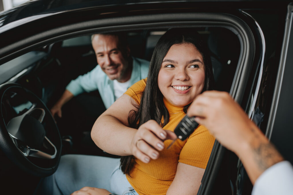 A young woman smiles as she receives car keys from someone outside the car, while a man sits beside her in the drivers seat, both looking happy.