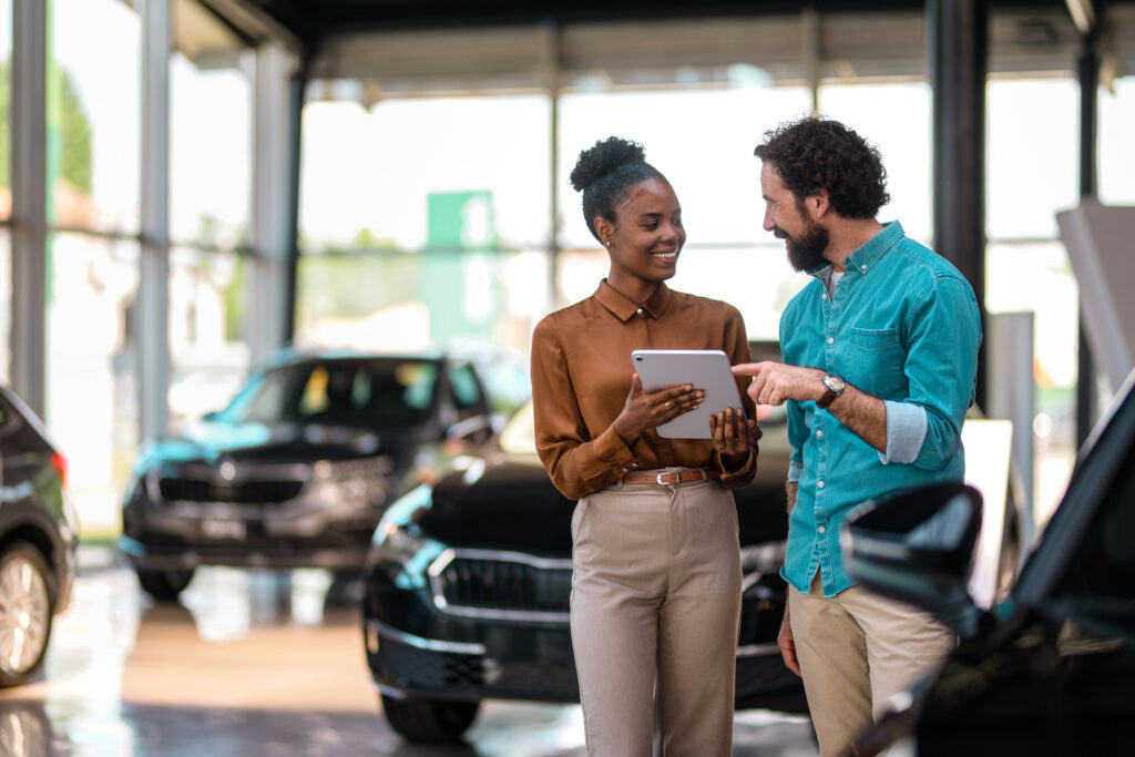 A woman holding a tablet and a man are smiling and talking inside a car dealership, surrounded by cars.