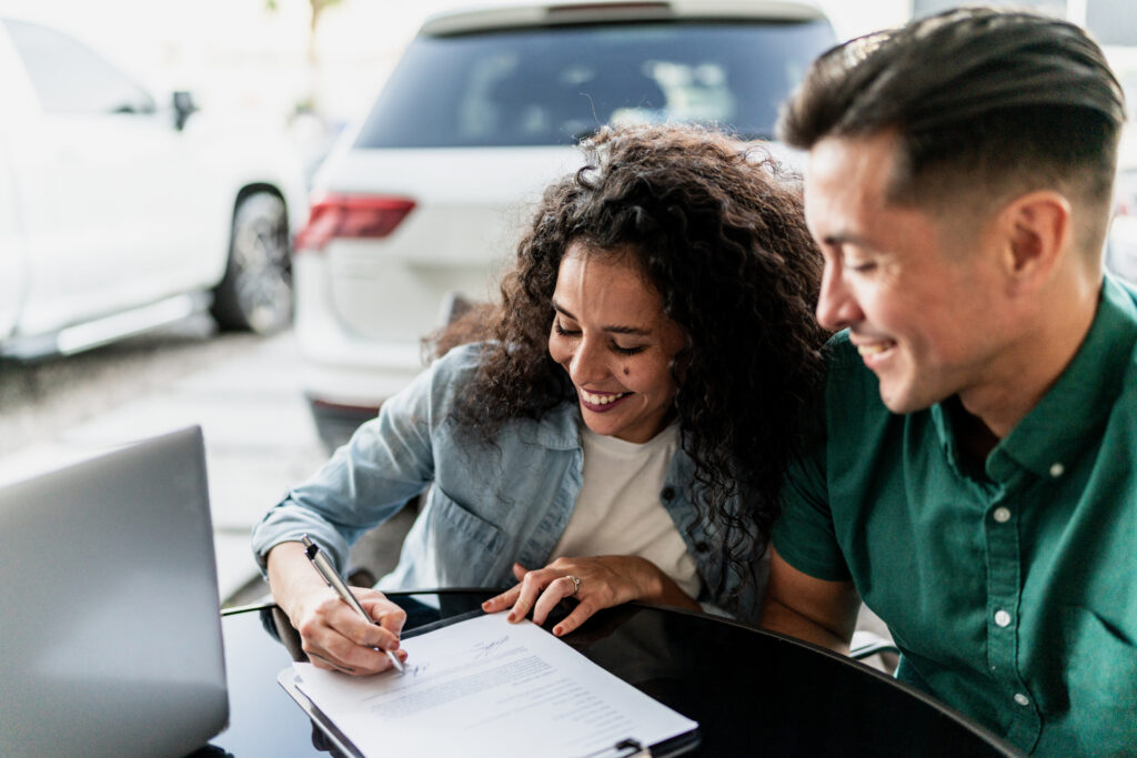 A woman smiles as she signs a document next to a man at a table outdoors, with a laptop and cars in the background.