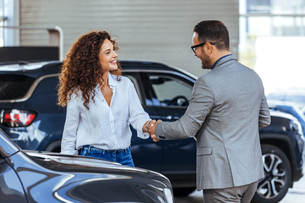 A woman smiling and shaking hands with a car salesman in a dealership.