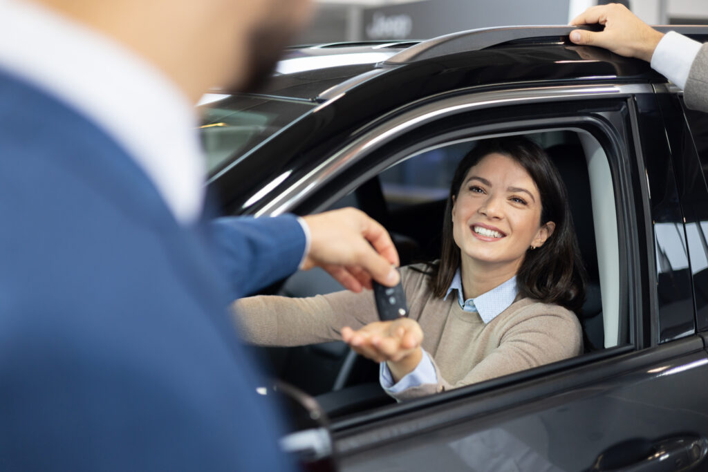A smiling woman sitting in the drivers seat of a car receives a key fob from a car salesperson.
