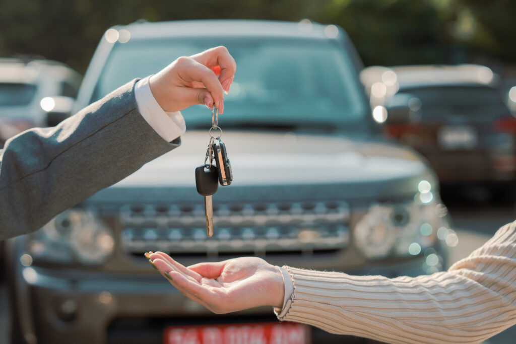 A person in a gray suit hands car keys to another person, with a vehicle in the background.